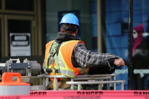 A construction worker wearing a blue helmet and safety vest operates equipment at an urban construction site.